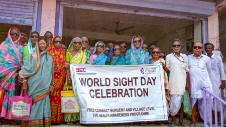 A group of people stand outside of a building holding a sign that reads "World Sight Day Celebration". All wear big, dark sunglasses.