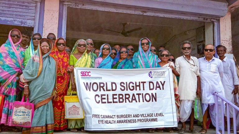 A group of people stand outside of a building holding a sign that reads "World Sight Day Celebration". All wear big, dark sunglasses.