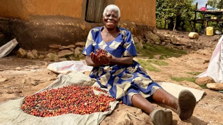 A woman sits next to a pile of raw coffee beans, smiling. She's holding up some of the beans in two hands.