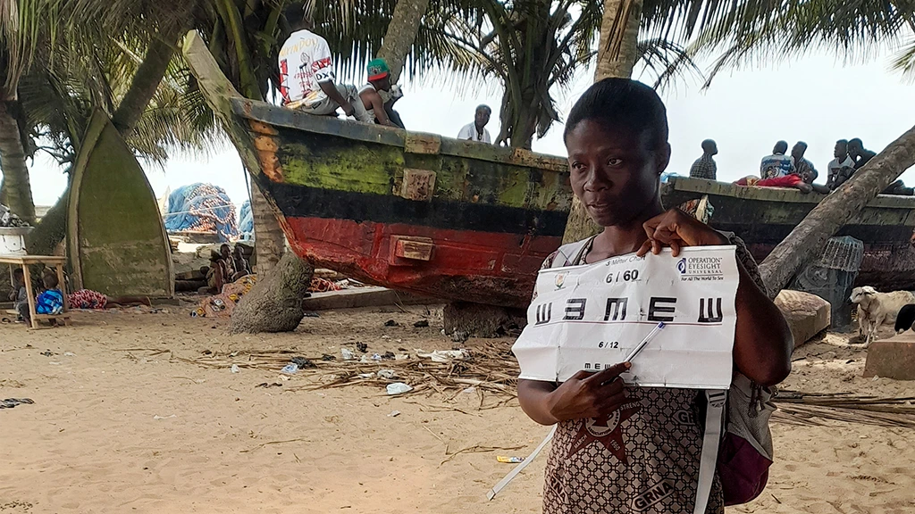 A woman holds up an eye chart. She is outdoors, and a boat and palm trees are visible in the background.