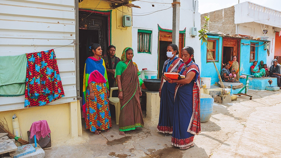 Three women stand outside their home talking to two community health workers in matching uniforms. One of the health workers takes notes on a clipboard.