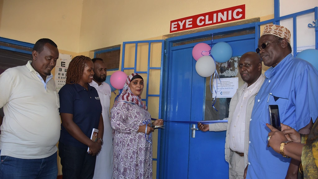 People stand next to a door with balloons afixed to it. A sign above the door reads "eye clinic". 