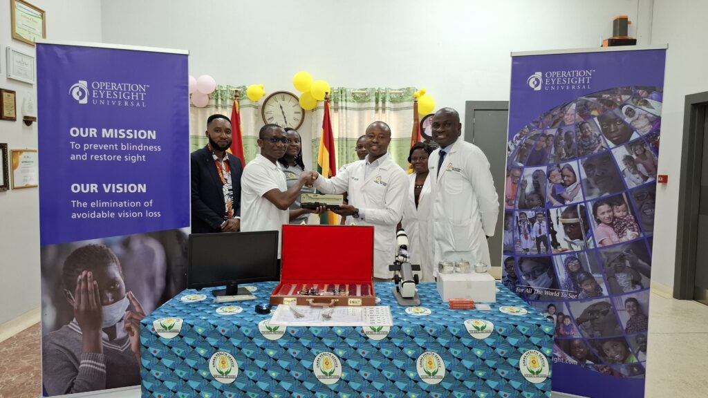 People stand between two banners with ophthalmic equipment laid out on a table in front of them. The two men in the centre are shaking hands.
