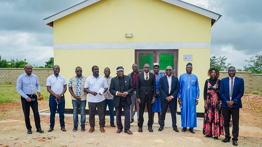 A group of men and women stand in a row in front of a small yellow building.