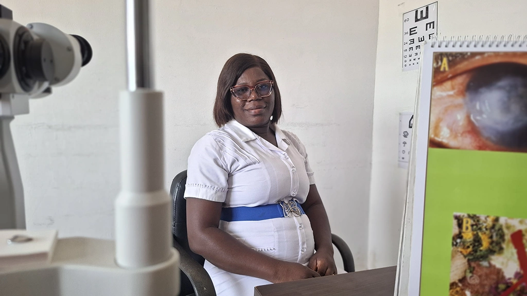 A woman poses from the camera in a nurses' uniform. She wears glasses and there is opthalmic equipment in the foreground. 
